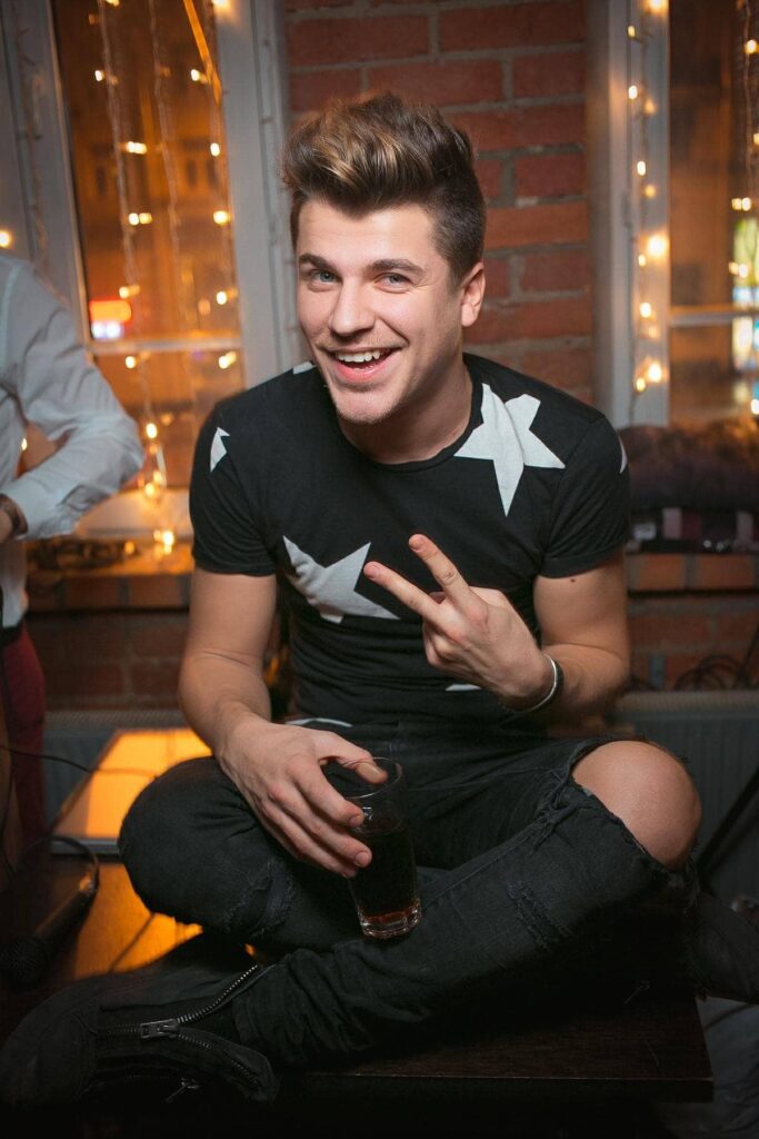 Парень с бокалом на вечеринке, улыбающийся и показывающий знак мира