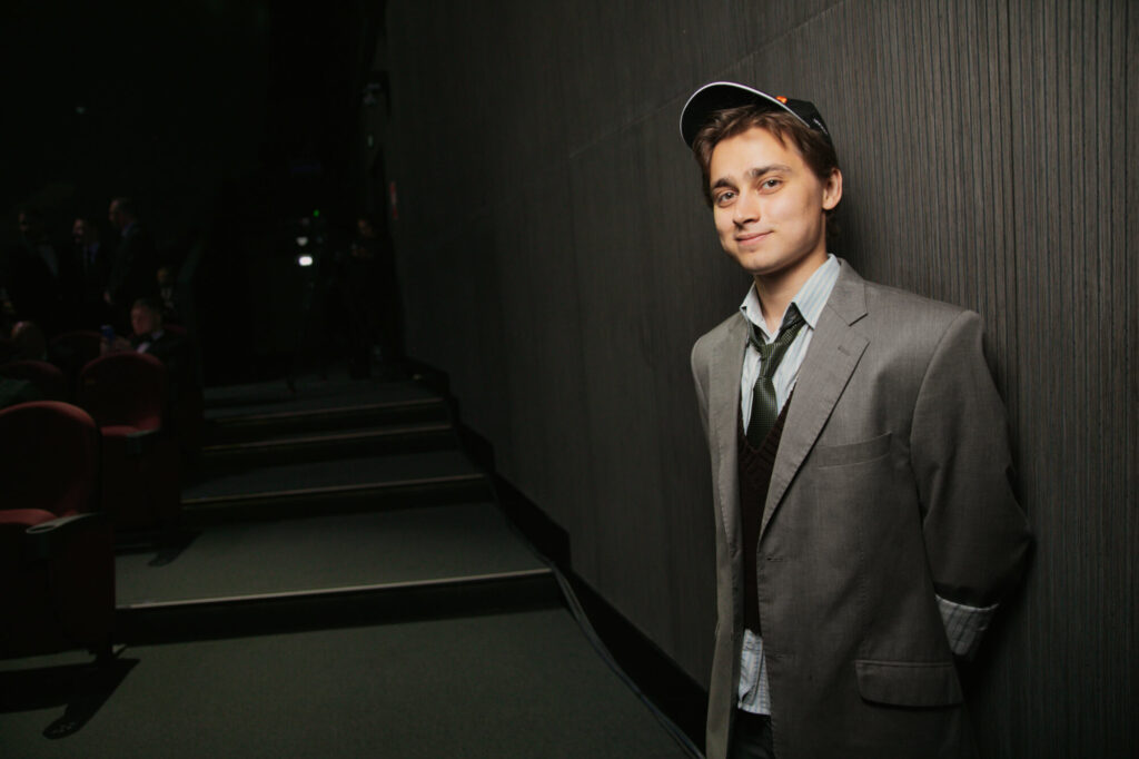 Юноша в пиджаке и кепке в расслабленной позе в темном помещении